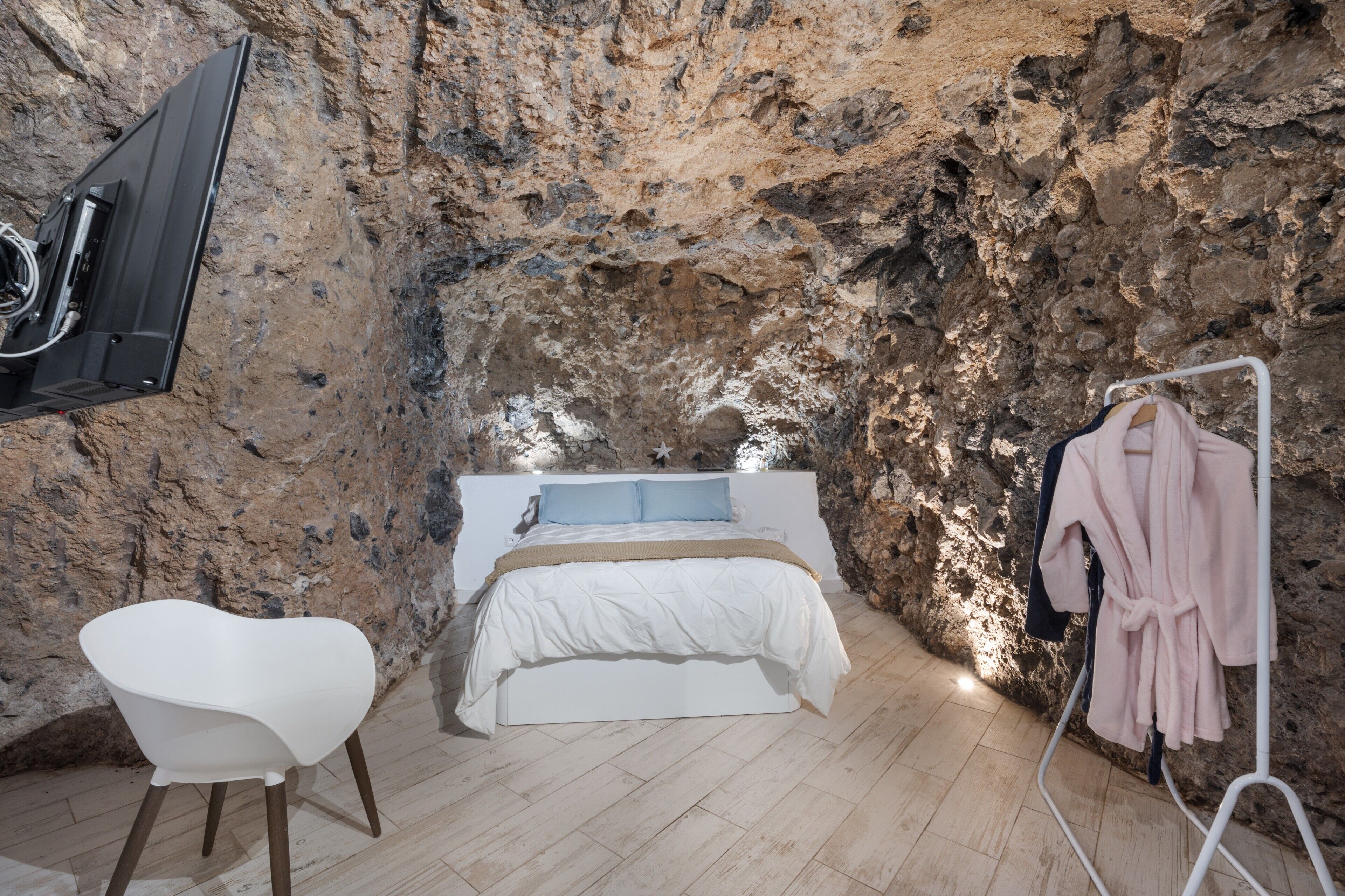 Ein Schlafzimmer mit einem Bett, einem weißen Stuhl, einem Kleiderständer und einer Steinwand.