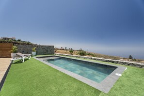 Une piscine avec un mur en pierre et un espace herbeux où se trouve une chaise.