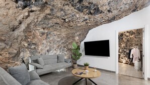 A modern living room with a grey sofa, a round coffee table, a TV mounted on the wall, and a large rock wall. - Luxury House 'La Cueva del Guanche' with Sea Views, Private Pool & Wi-Fi (Adeje)