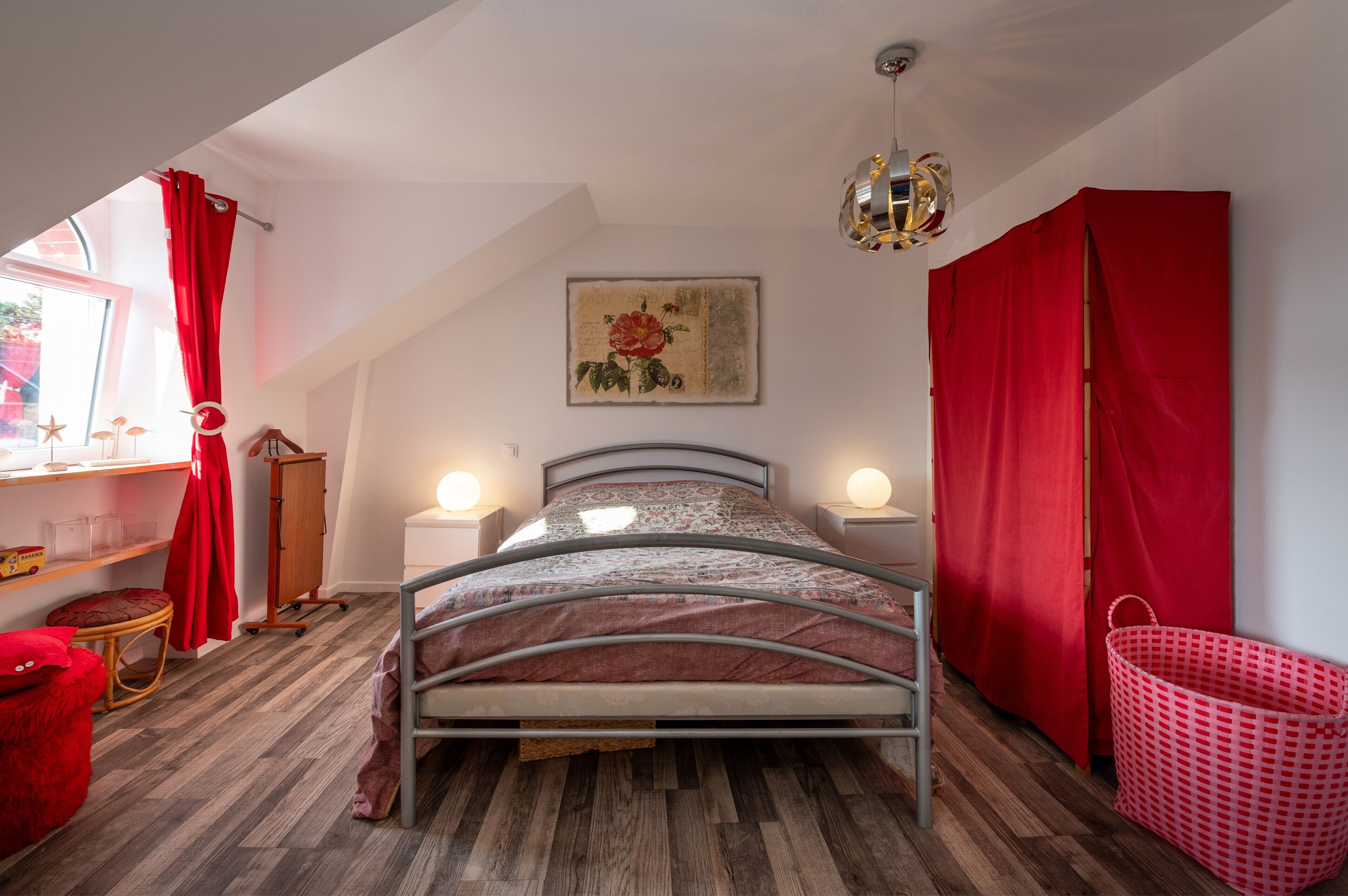 A bedroom with a metal bed frame, wooden flooring, a red curtain, a wall-mounted lamp, and a red basket.