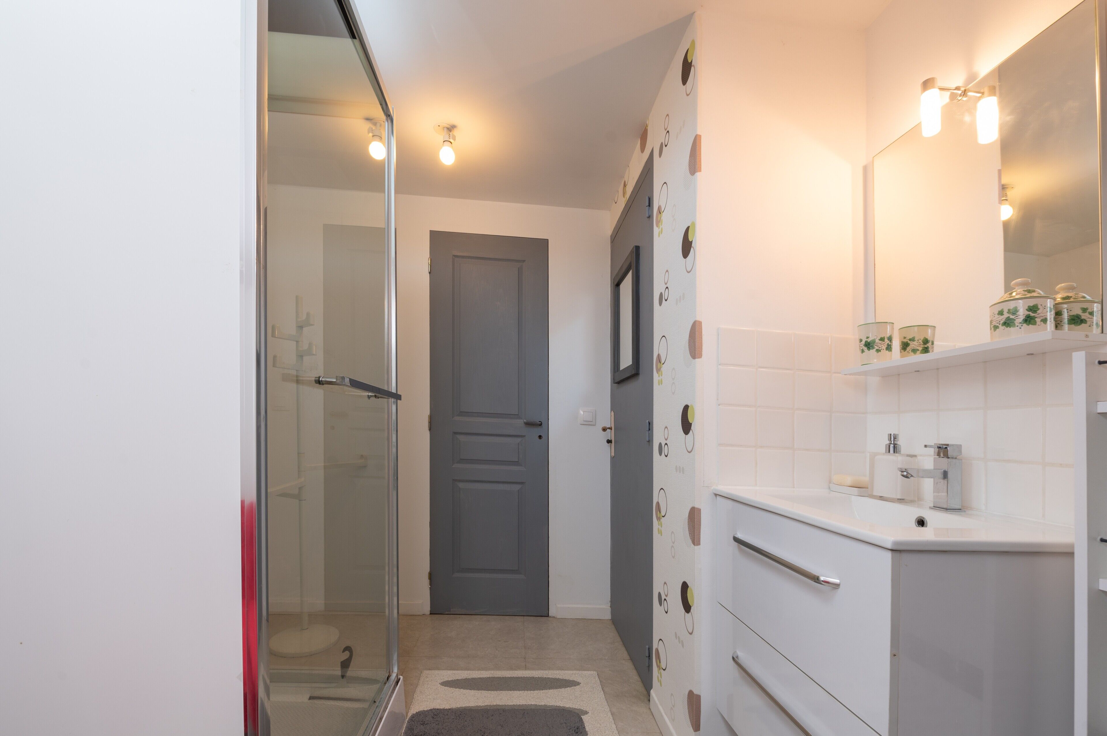 A bathroom with a glass shower enclosure, a white sink with a mirror, and a door leading to another room.