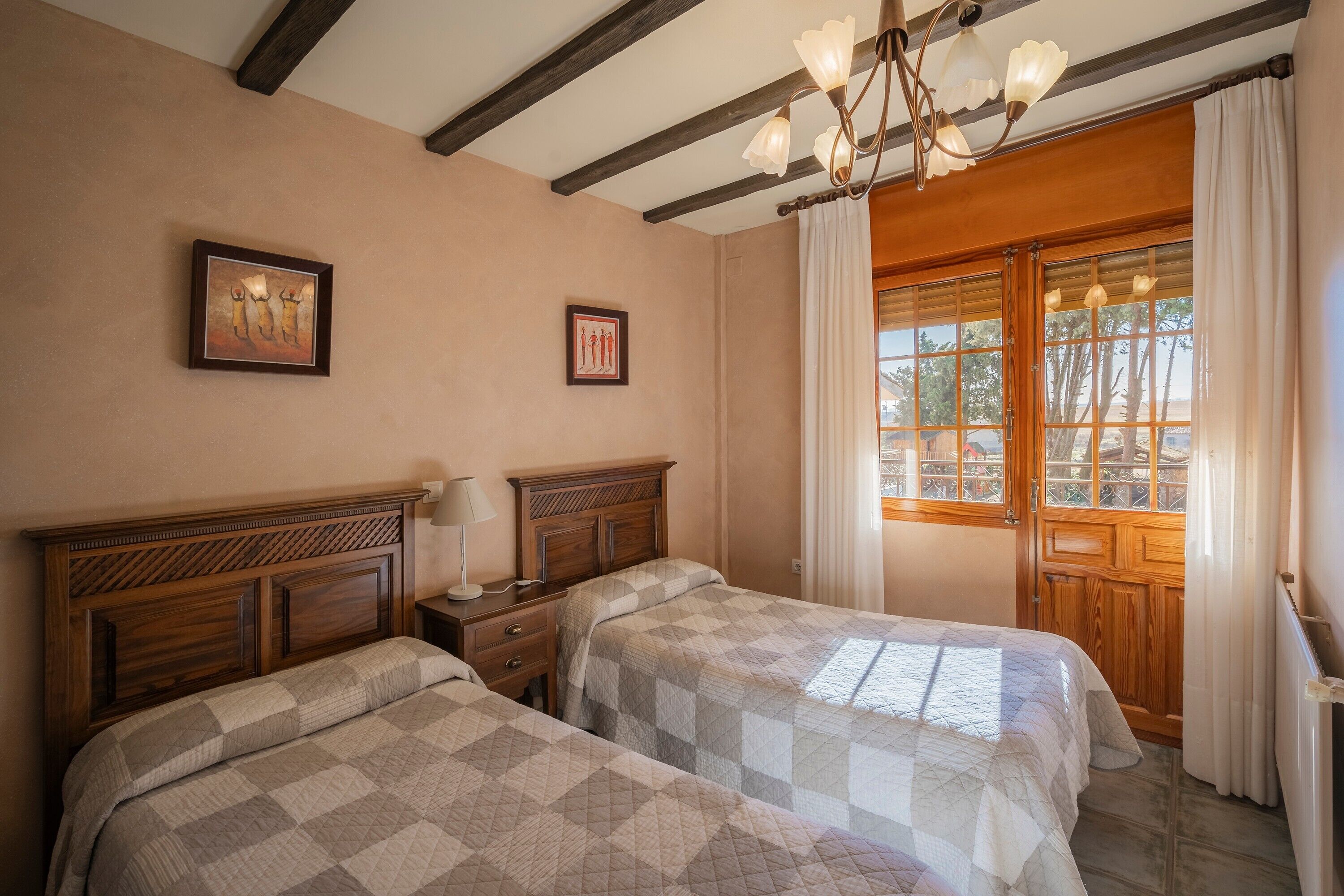 Une chambre à coucher avec deux lits, des têtes de lit en bois, un lustre, une fenêtre avec des rideaux et des tableaux encadrés accrochés au mur.