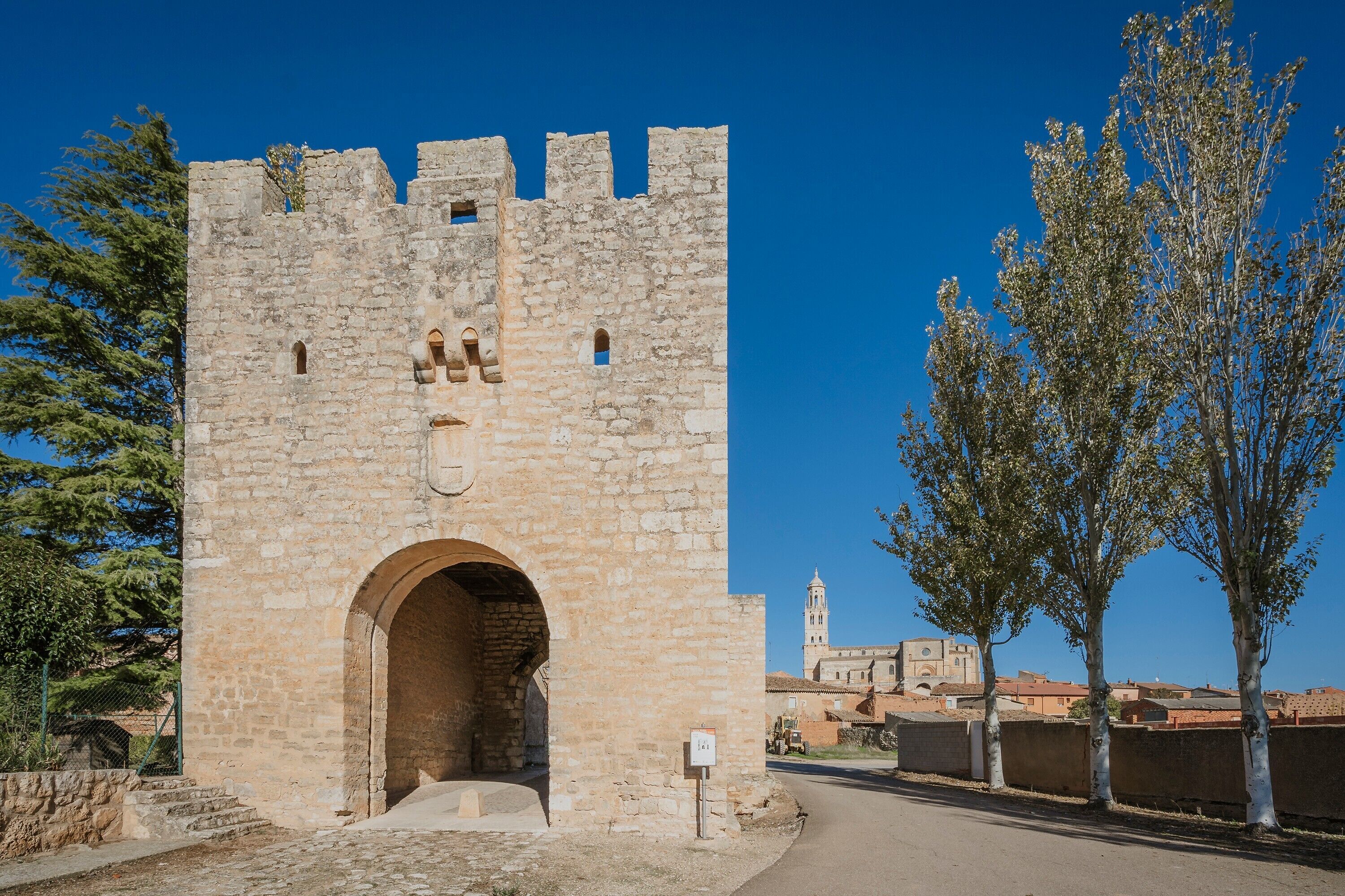 Un castillo de piedra con un gran arco de entrada, cielo despejado e iglesia a lo lejos.