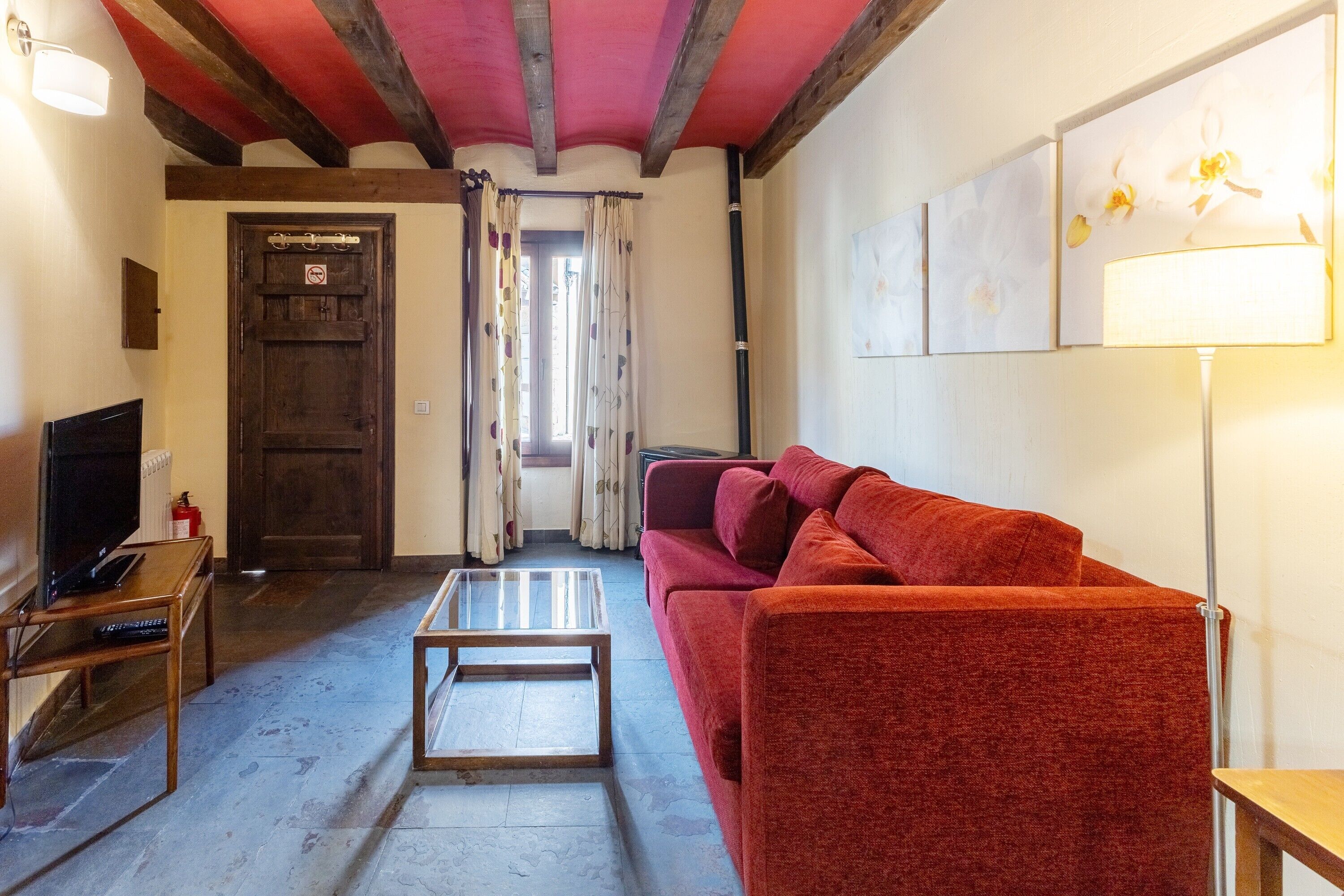 Un salon avec un plancher en bois, un canapé rouge, une table basse en verre, un téléviseur posé sur un meuble en bois et une porte avec un rideau.