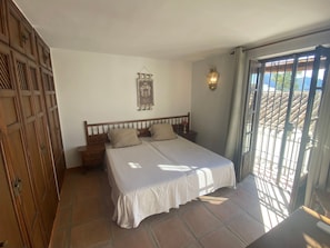 Une chambre à coucher comprenant un lit, une table de chevet, une lampe fixée au mur, un élément décoratif suspendu et une vue sur l’extérieur, à travers une fenêtre ornée d’une grille en métal.