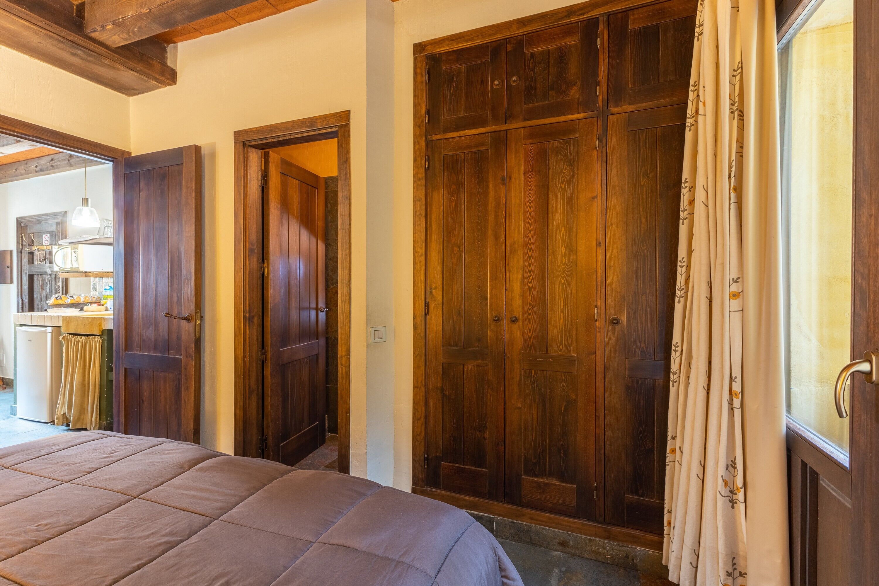 Une chambre avec un grand lit, une armoire en bois et une cuisine visible à travers une porte ouverte.