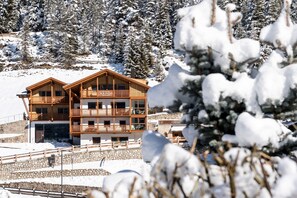 Un immeuble en bois à plusieurs étages, doté de balcons, entouré d’arbres couverts de neige et d’un mur de pierre.