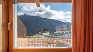 A room with a large window overlooking a mountainous landscape, a wooden balcony, and a view of a house and green fields. - Apartment 'Villa Solinda App Vivaldi' with Mountain View, Private Terrace and Wi-Fi (Selva)