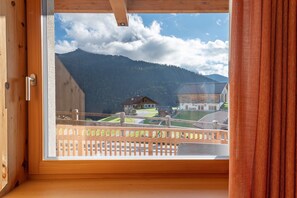 Une pièce dotée d’une grande fenêtre donnant sur un paysage montagneux, d’un balcon en bois et d’une vue sur une maison et des champs verdoyants.