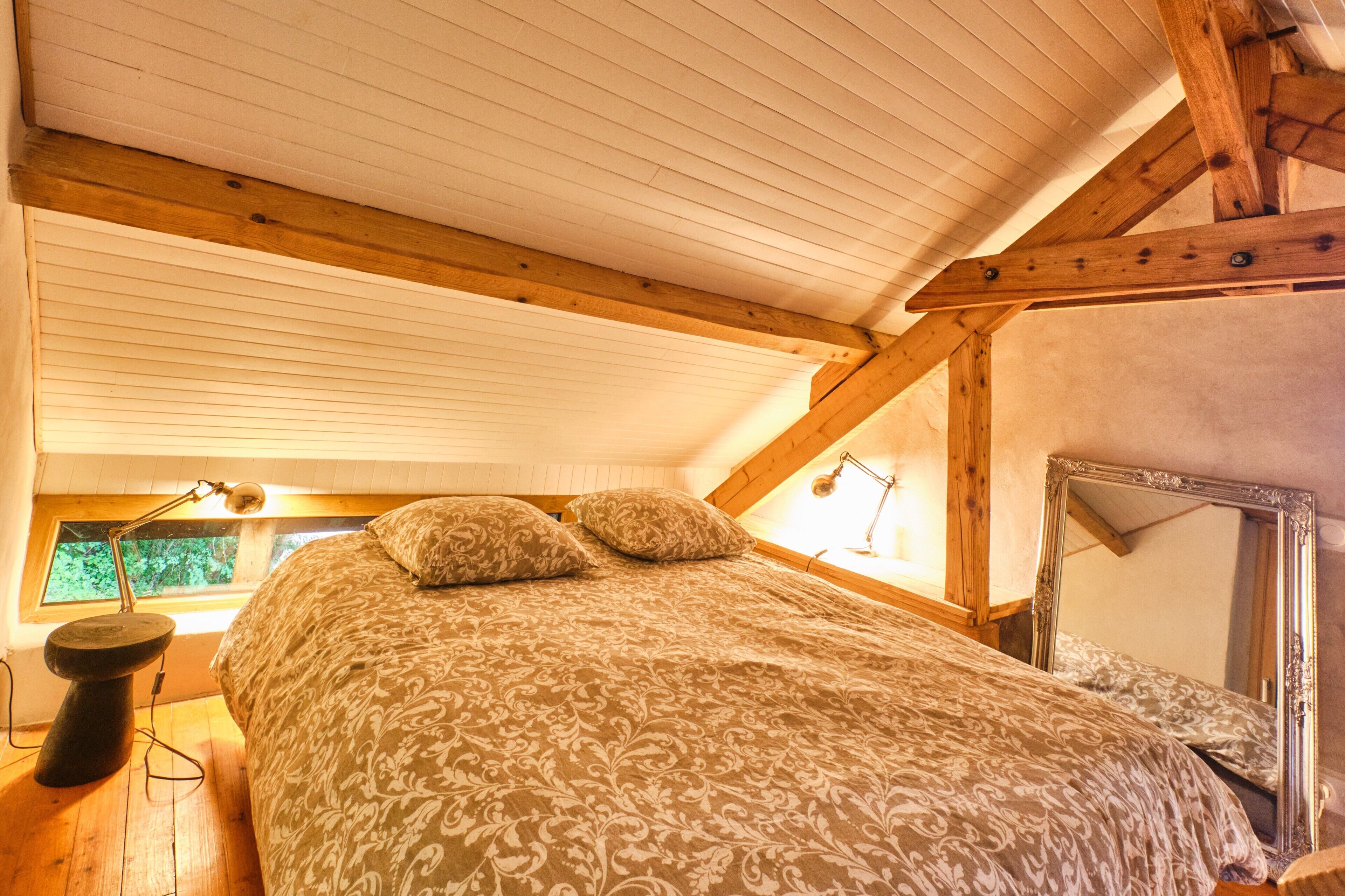 Un dormitorio con techo inclinado, cama con ropa de cama estampada, lámpara de noche, espejo y ventana con vistas a follaje.
