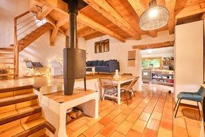 Ein rustikaler Innenraum mit einer Holztreppe, einem zentralen Kamin, einem Essbereich und einer durch einen offenen Durchgang sichtbaren Küche.