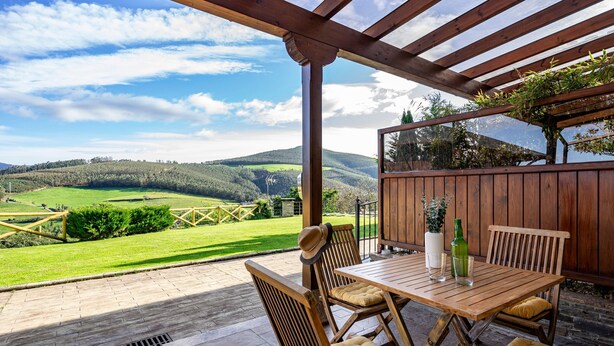 A wooden pergola with a table and chairs, a view of a green landscape, and a wooden fence.