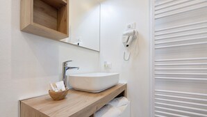 Un baño moderno con tocador de madera, un lavamanos blanco, un secador de pelo en la pared y un perchero para toallas.