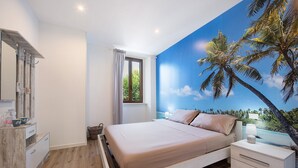 Un dormitorio con cama, mesita de noche, cómoda, ventana con cortinas y un mural de palmeras.