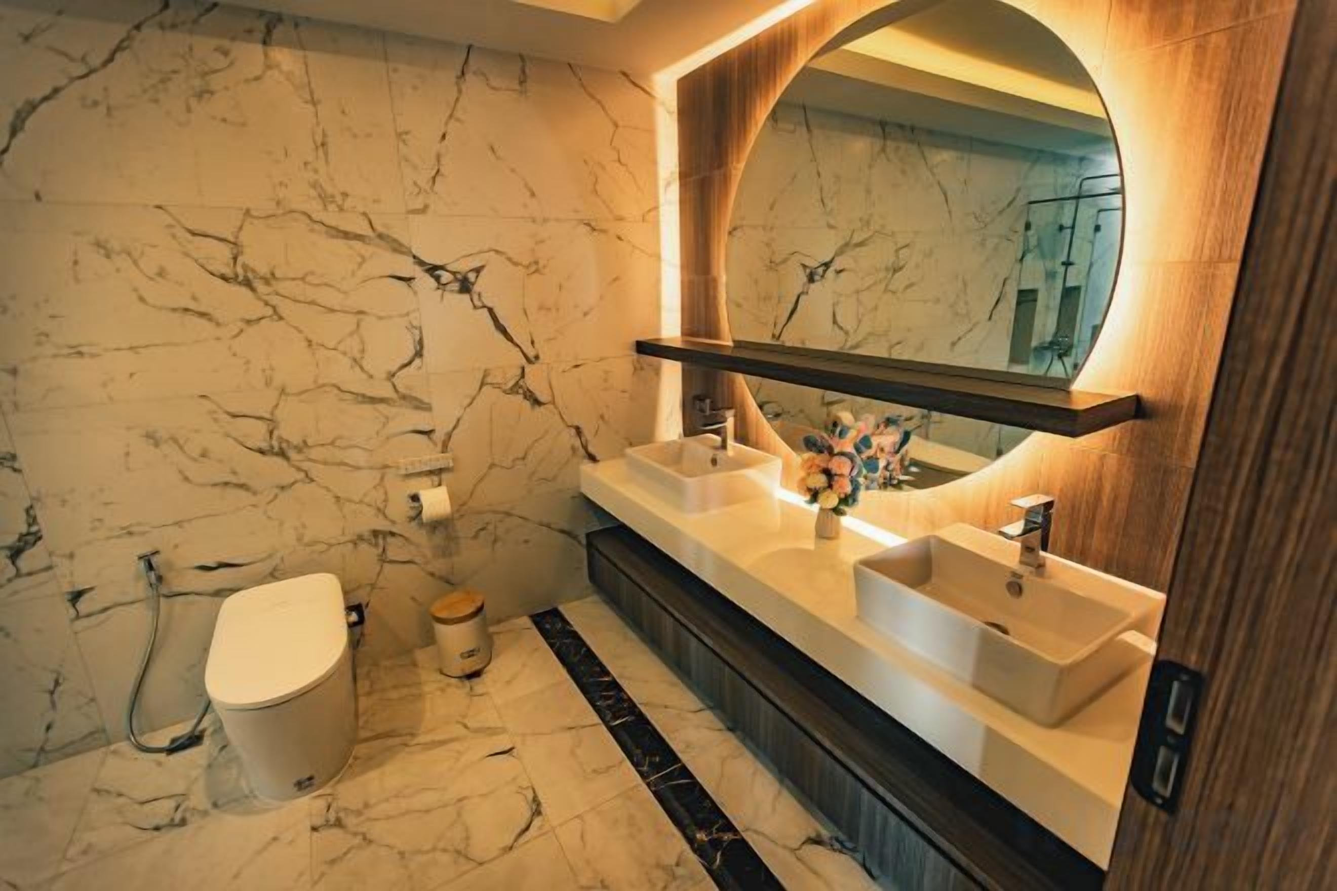 Une salle de bain aux murs recouverts de marbre, avec des toilettes blanches et un meuble-lavabo doté de deux vasques et d’un miroir.
