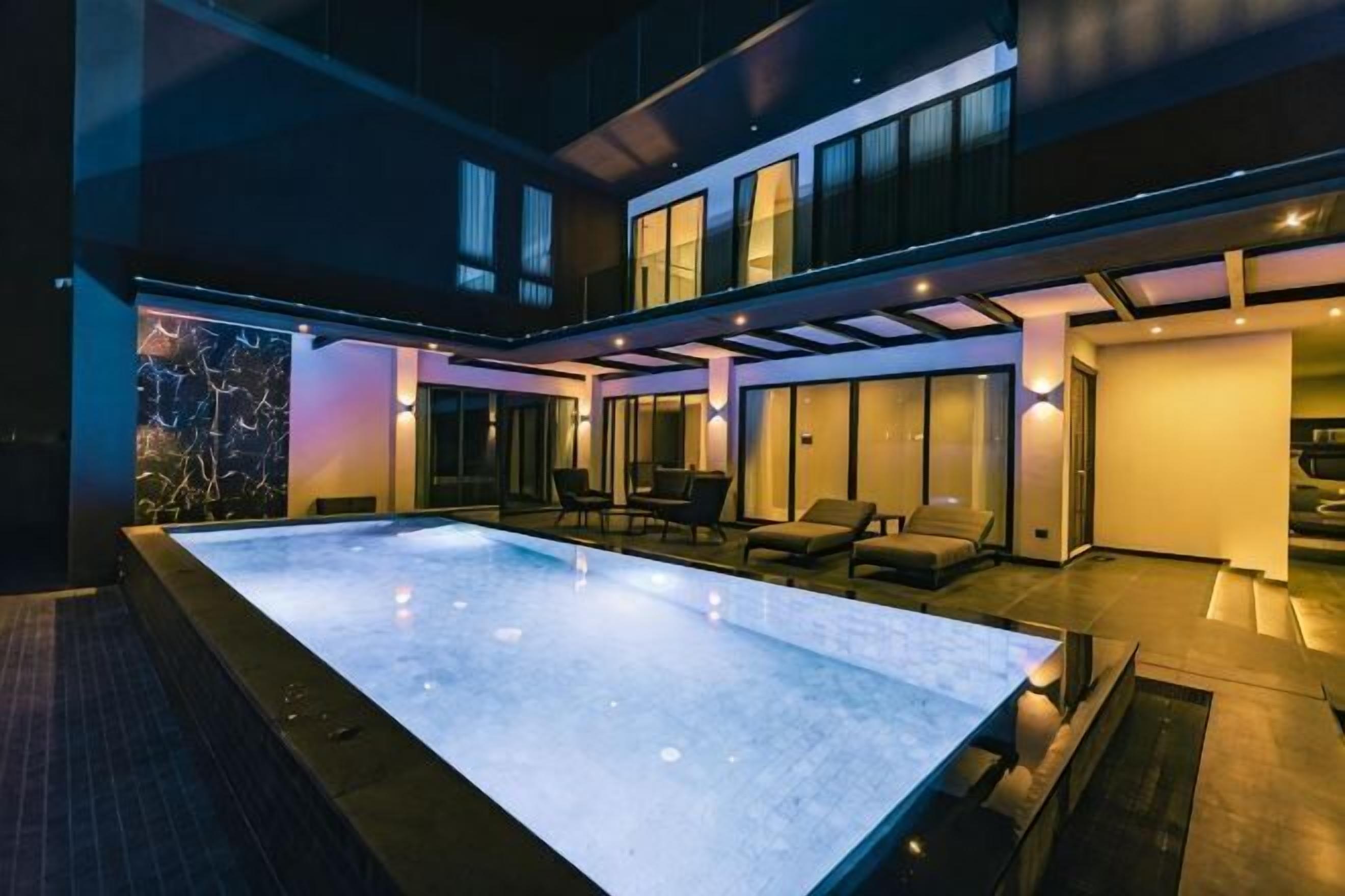 A modern outdoor swimming pool area with a glass enclosure, seating, and a well-lit interior space.