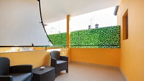 A balcony with two wicker chairs and a small table, a yellow wall, and a green hedge in the background.