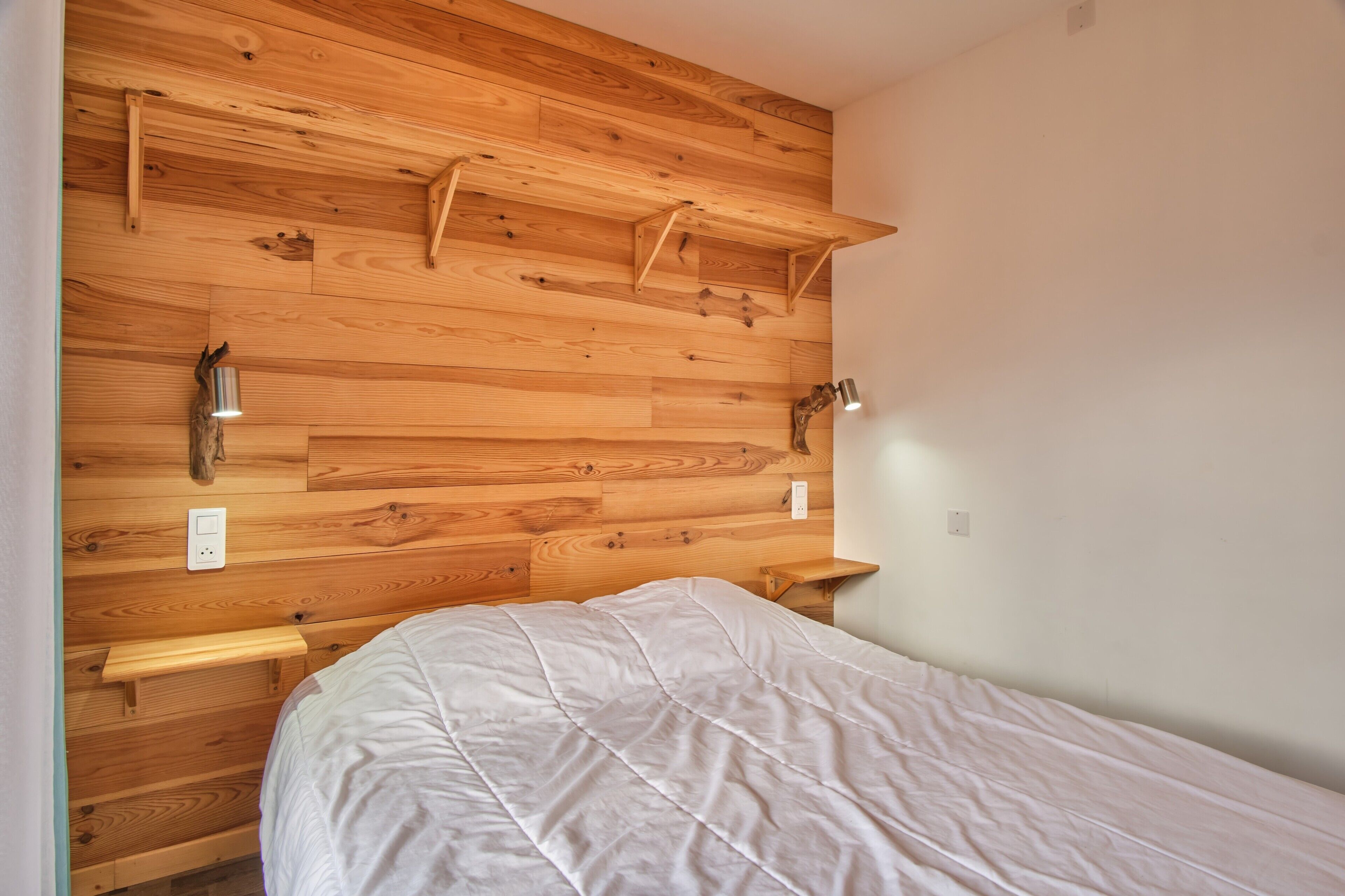 Une chambre avec une tête de lit en bois, un lit recouvert d’une couette blanche, des tables de chevet et des lampes fixées au mur.