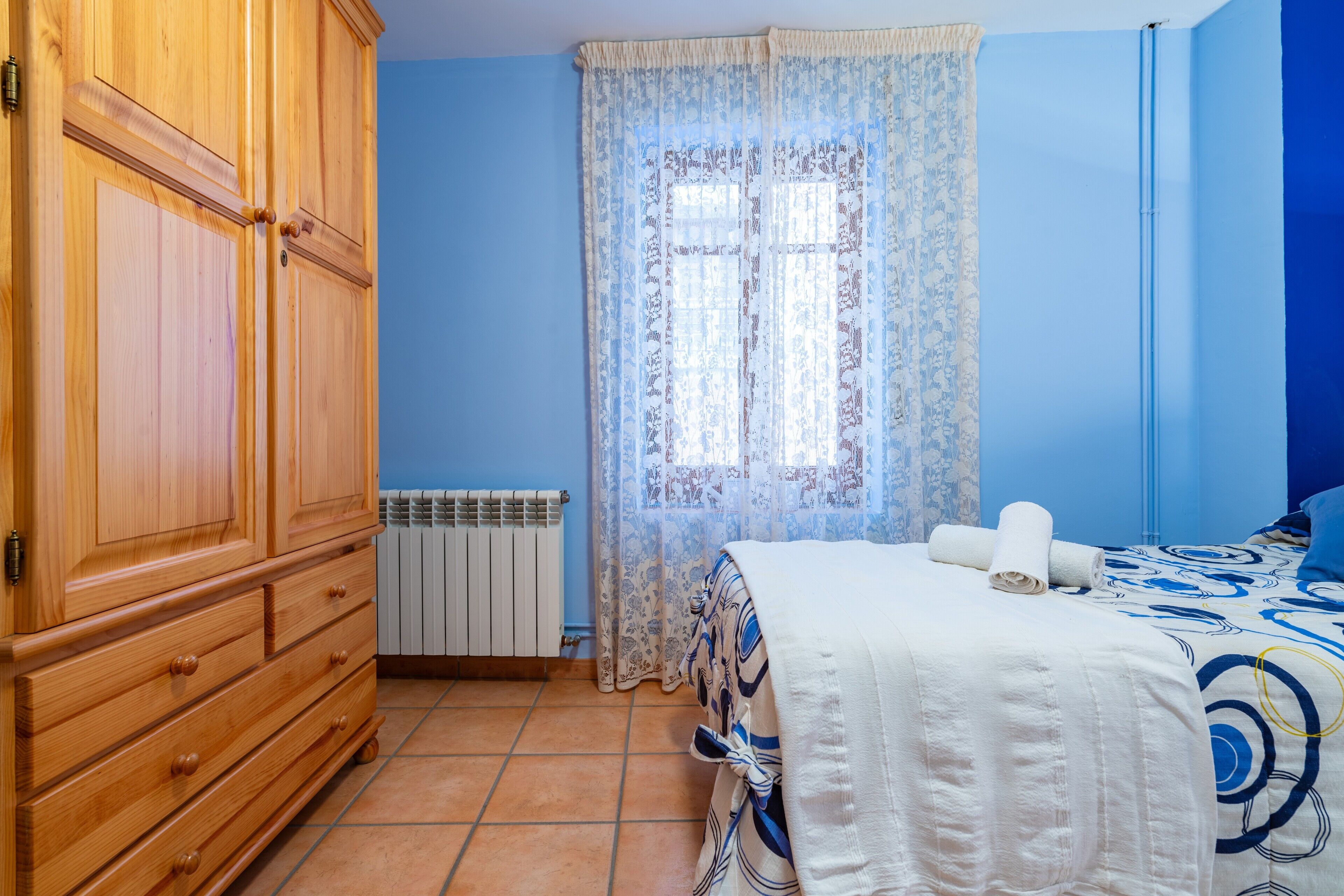 A bedroom with a wooden wardrobe, a radiator, a window with patterned curtains, and a bed with a blue and white bedspread.