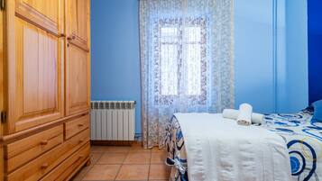 A bedroom with a wooden wardrobe, a radiator, a window with patterned curtains, and a bed with a blue and white bedspread.
