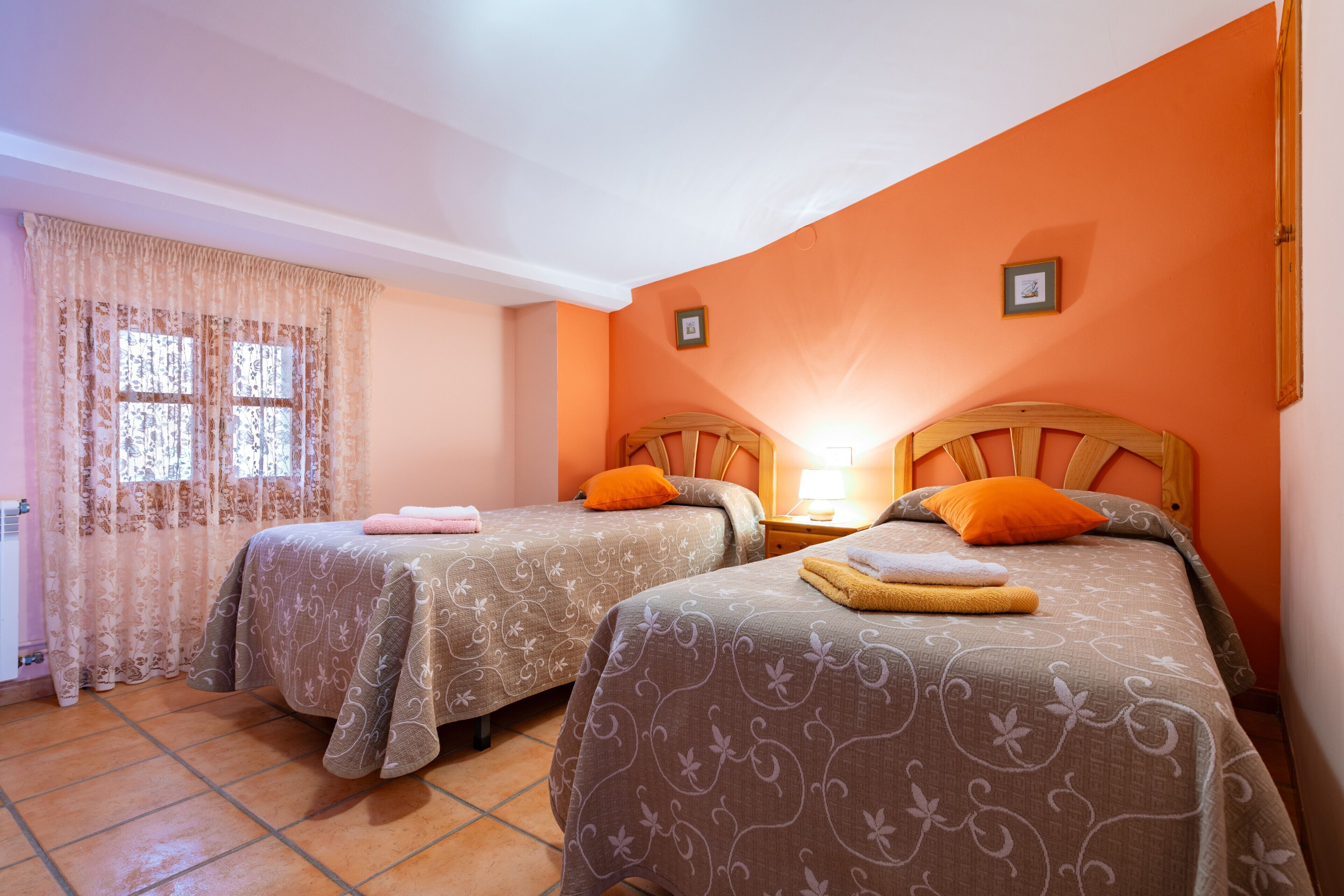 Una habitación con dos camas, una ventana con cortinas, un cabecero de madera y una mesita de noche con lámpara.