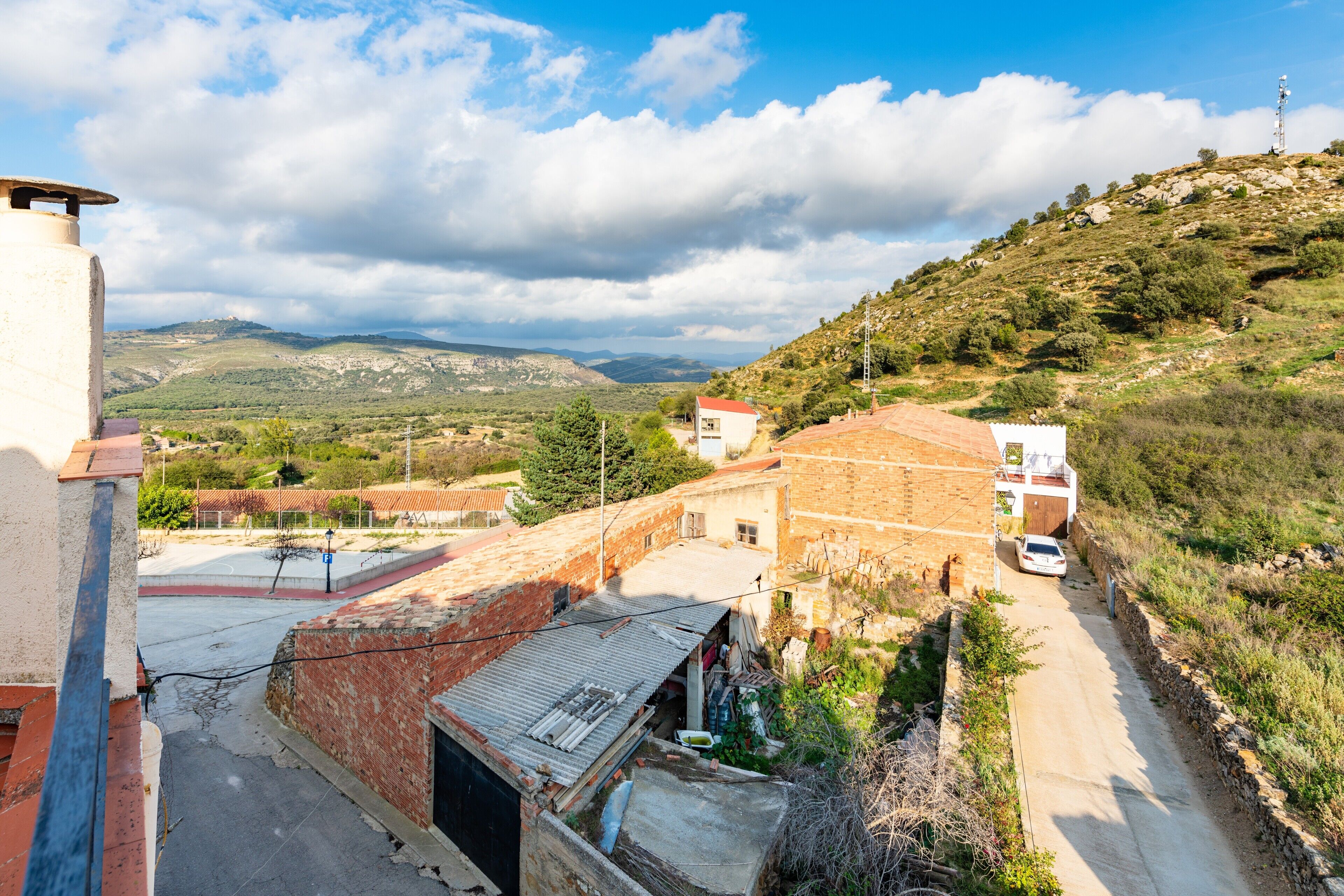 Una carretera rural con un coche blanco, edificios con techos rojos y un paisaje montañoso al fondo.