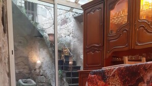 Een kamer met een stenen vloer, een tafel met een notentop en fruitprint, een houten kast en uitzicht door een glazen deur op een buitenruimte met een stenen muur en een metalen poort.