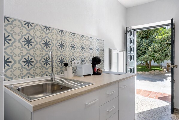 Una cocina con un lavamanos de acero inoxidable, armarios blancos, un salpicadero decorado y vistas a un jardín a través de una puerta abierta.