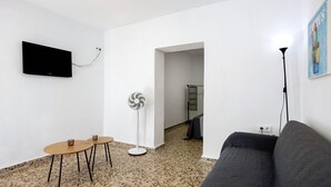 A living room with a flat-screen TV, two round tables, a standing fan, a grey sofa, and a cactus wall art piece. - Cozy Vacation Rental in Chiclana de la Frontera (Chiclana de la Frontera)