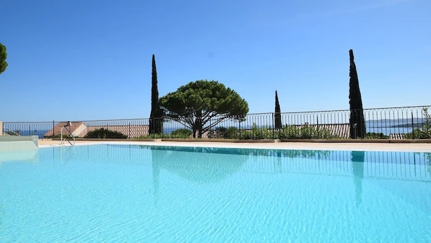 Una piscina azul clara con una valla metálica, un árbol y vista al mar.