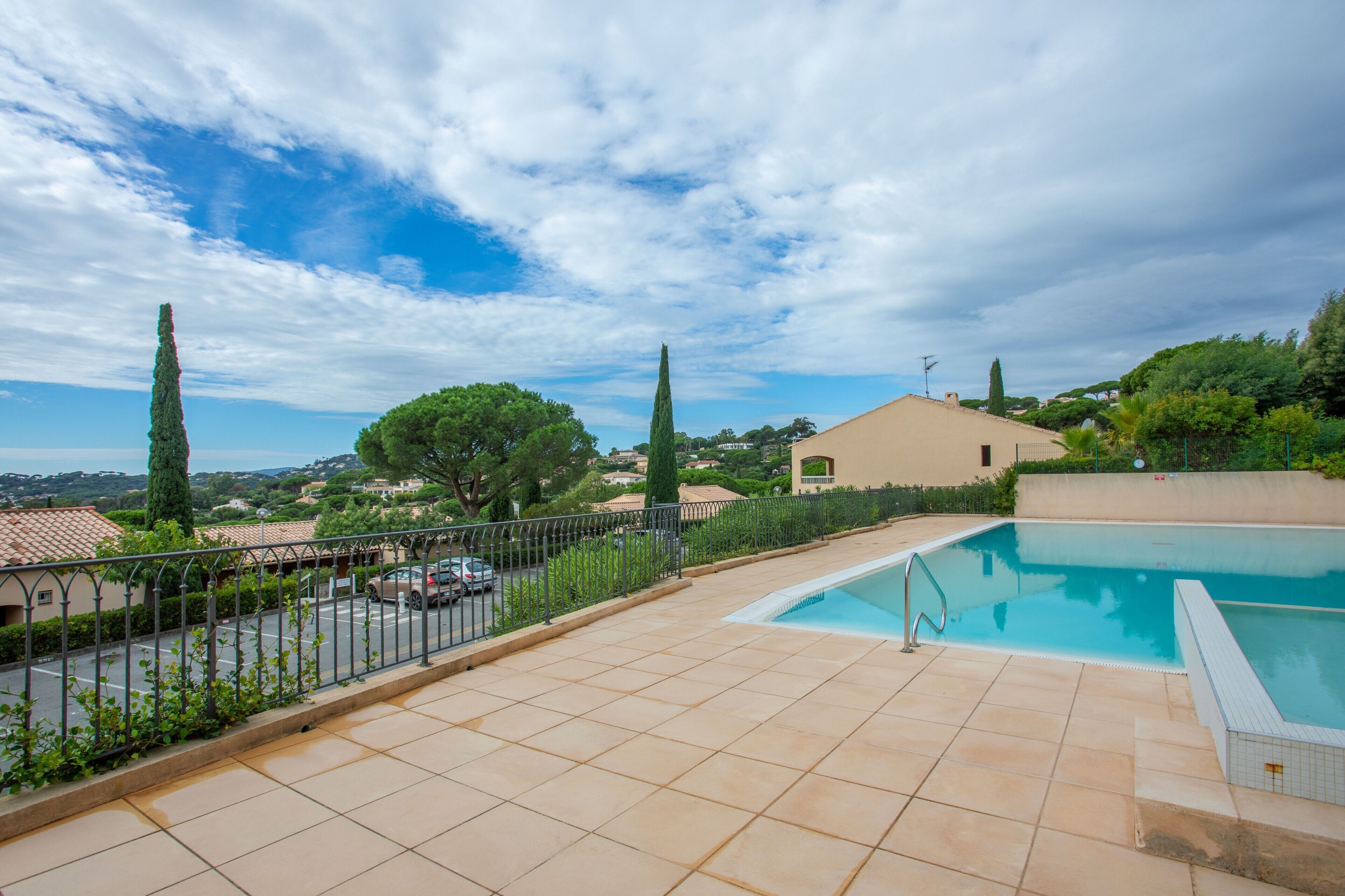 A swimming pool with a tiled deck, a fence, and a view of a residential area with houses and trees.