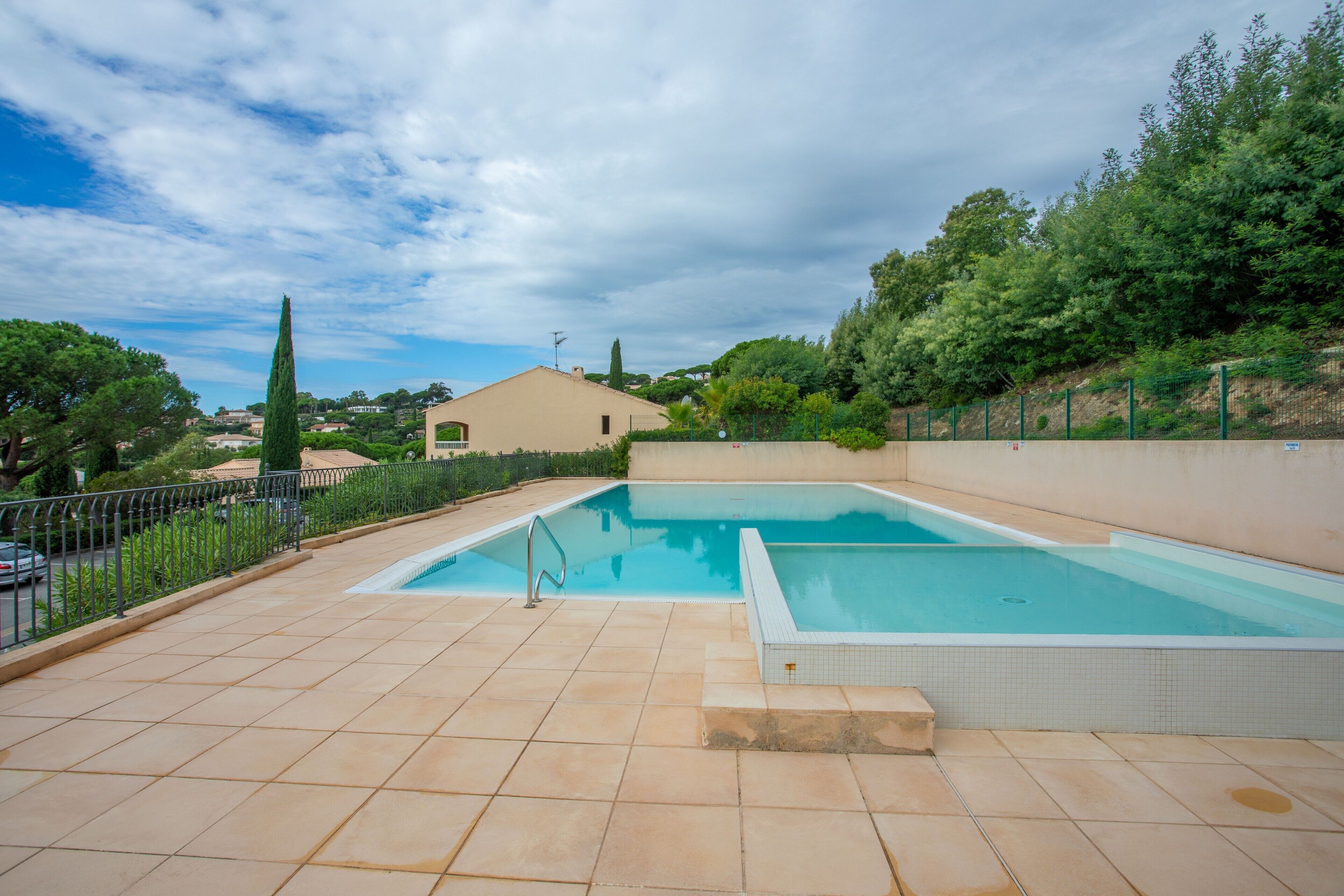 A rectangular swimming pool with a tiled deck, surrounded by a fence and greenery.