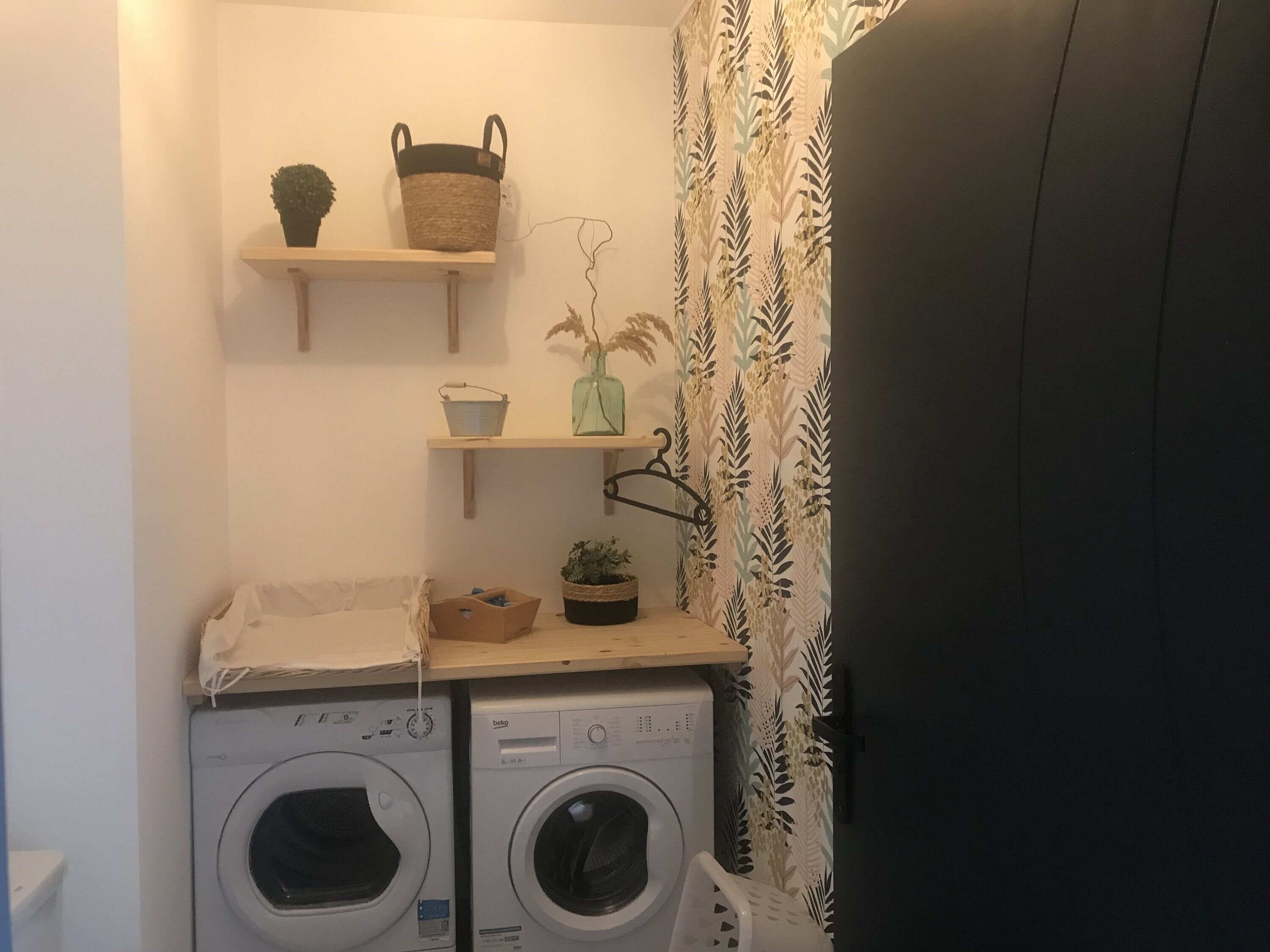 A laundry area with two washing machines, shelves with baskets and plants, and a patterned curtain.