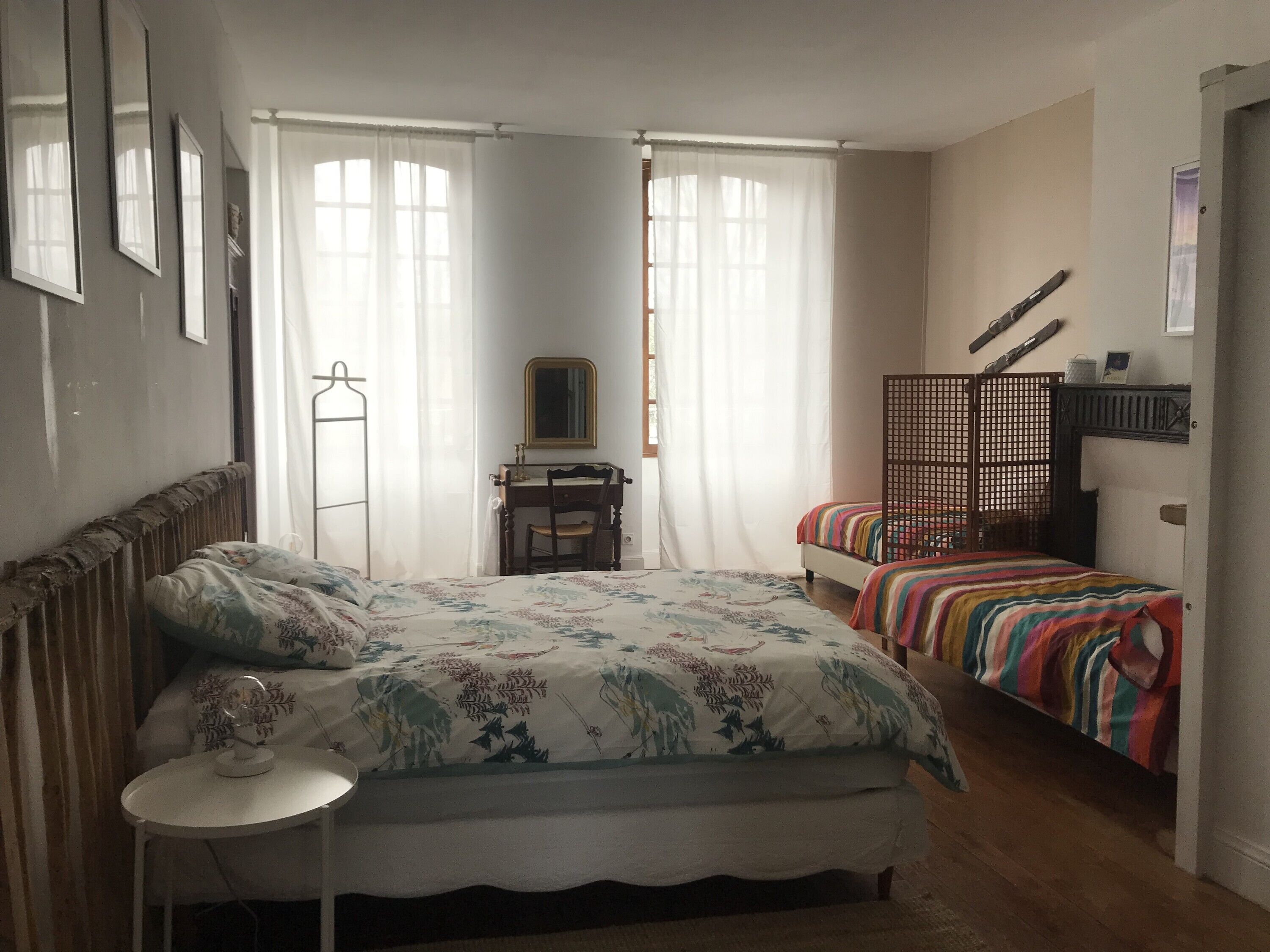 A bedroom with a bed, bedside table, and a chair. There is a desk with a chair and a mirror. The room has two windows with curtains.