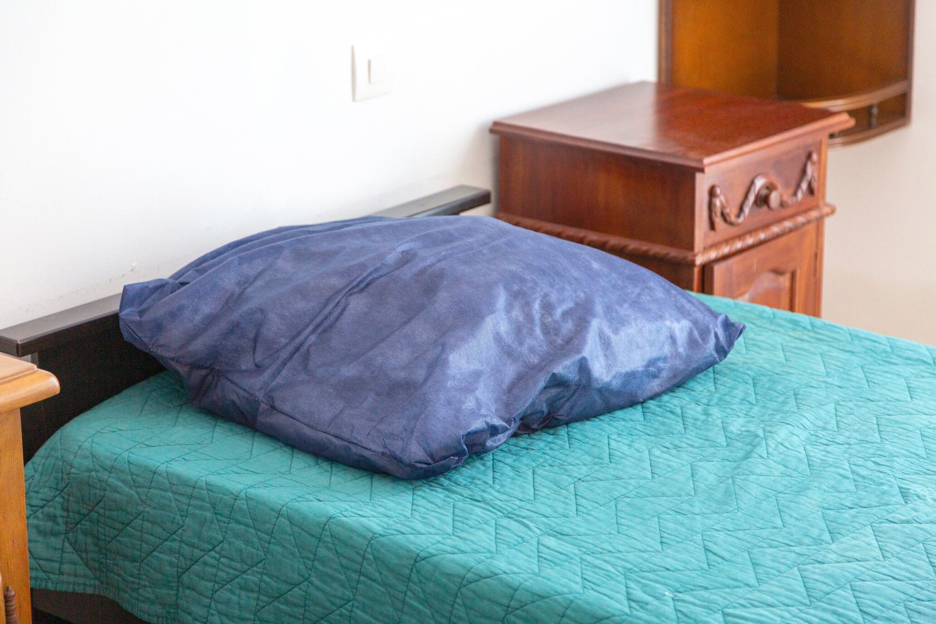 Un lit recouvert d’une courtepointe turquoise et orné d’un coussin bleu, dans une pièce où l’on trouve une table de chevet en bois et un téléviseur.