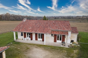 Une maison de plain-pied avec un toit recouvert de tuiles rouges, une véranda abritée avec des chaises blanches et un petit jardin.