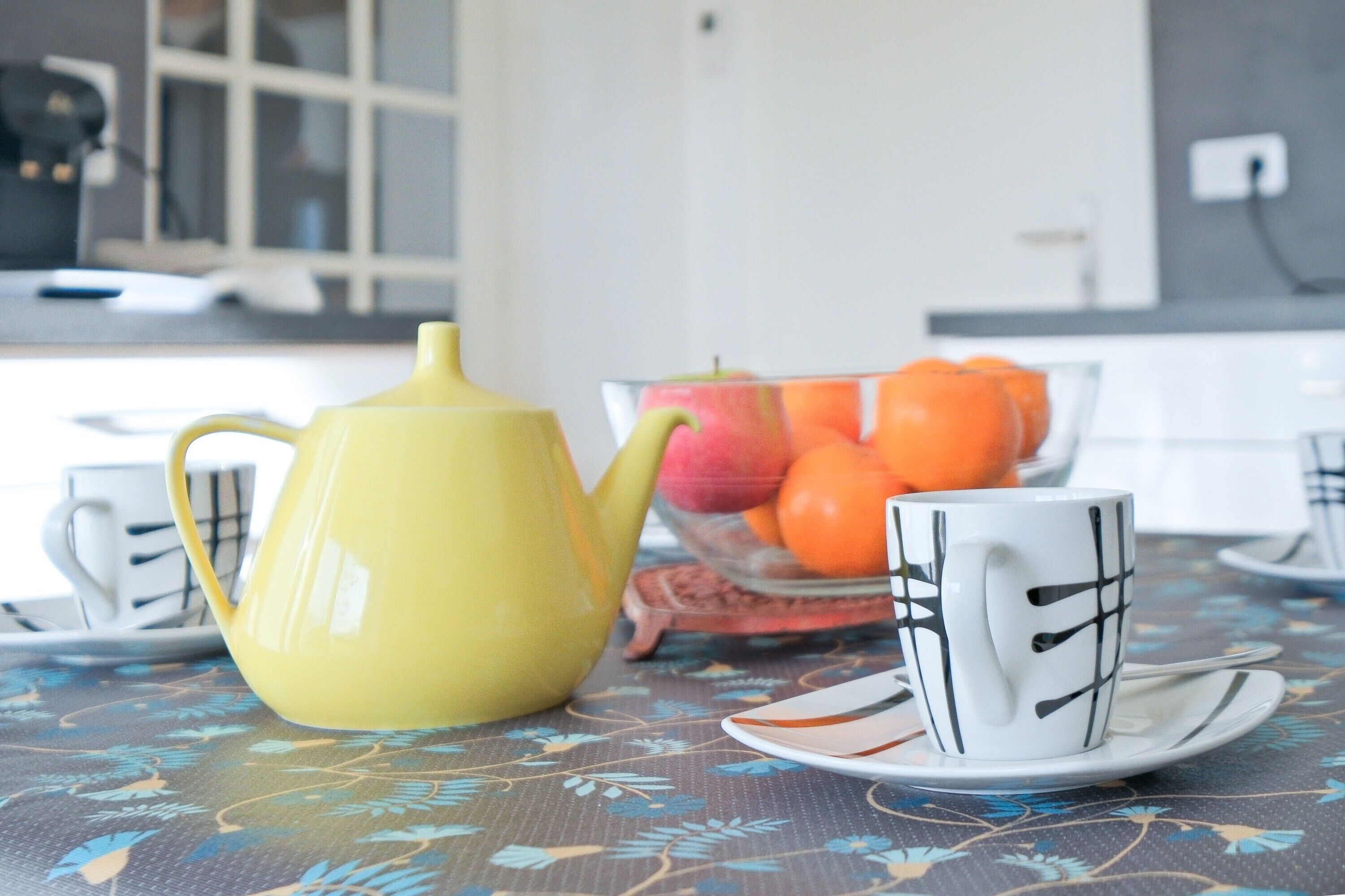Eine Küche mit einem gelben Teekessel, einer Fruchtschale und einer weißen Tasse mit schwarzem Muster auf einem Tisch.