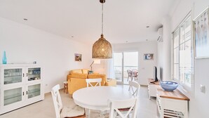 Un espace repas comprenant une table ronde, des chaises blanches, un luminaire suspendu, un canapé et un meuble avec des portes en verre.