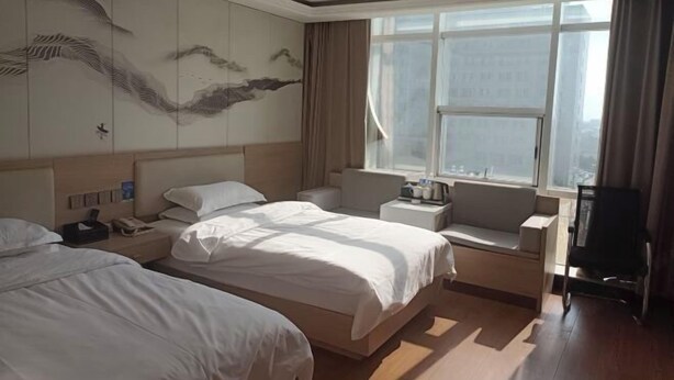 Ein Hotelzimmer mit zwei Betten, einem großen Fenster, einer Wand mit Kunstwerken und Blick auf die Stadt.