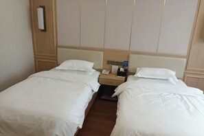 A hotel room with two single beds, a wooden headboard, a nightstand with a phone and a bottle, and a wall-mounted radiator.