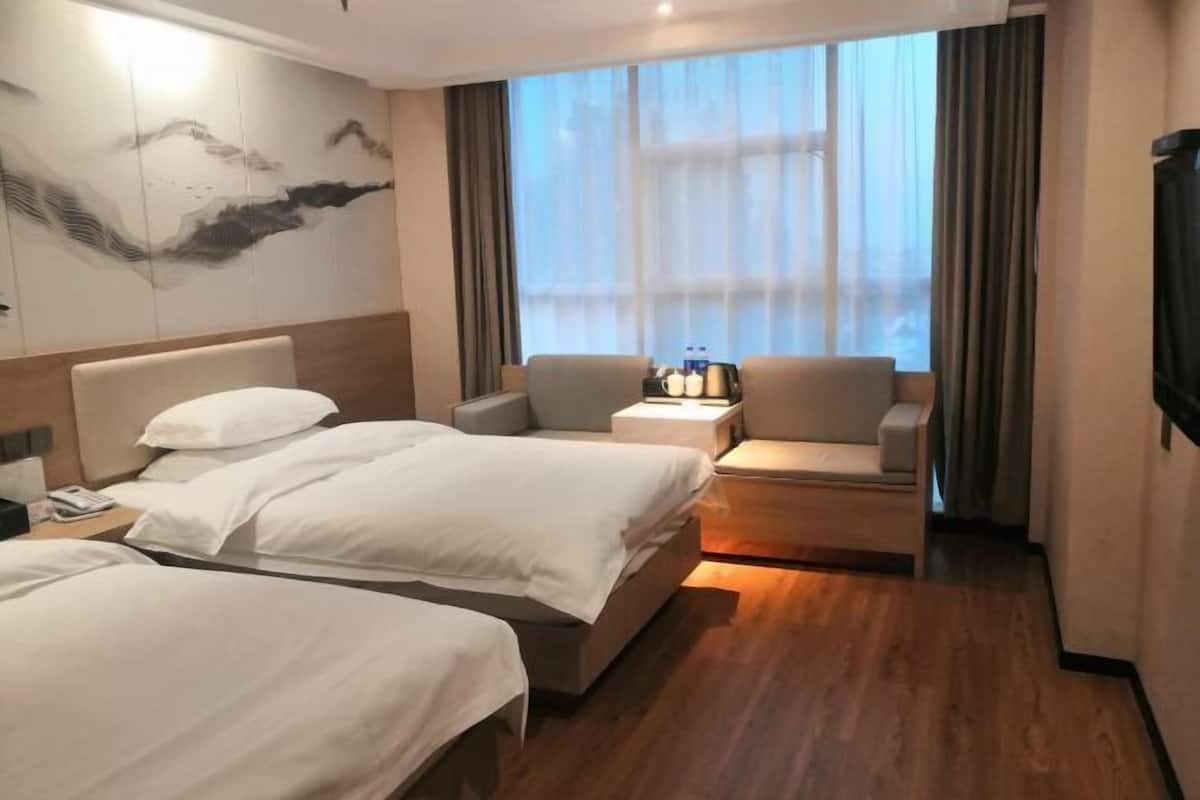 A hotel room with two beds, a sofa, a small table with bottles, and a large window with curtains.