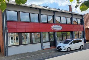 Rakennuksessa on punainen markiisi, sen edessä on valkoinen auto ja taivas on kirkas.