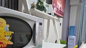 A microwave oven with a digital display and control panel, a small plant on a shelf, and a television in the background. - 1 Bedroom Studio Sleeps 2 -Elevator -Free Parking (Nairobi)