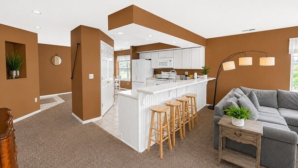 Une cuisine moderne avec des meubles blancs, un coin bar avec des tabourets, et un canapé gris dans un salon aux murs et au tapis bruns.