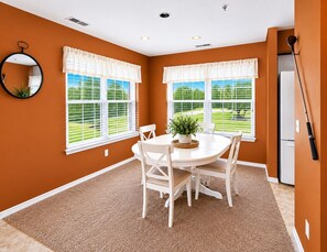 A dining area with a white table and chairs, two windows with white blinds, a potted plant on the table, and a kitchen area with a refrigerator. - Lounging on the Links (Branson)
