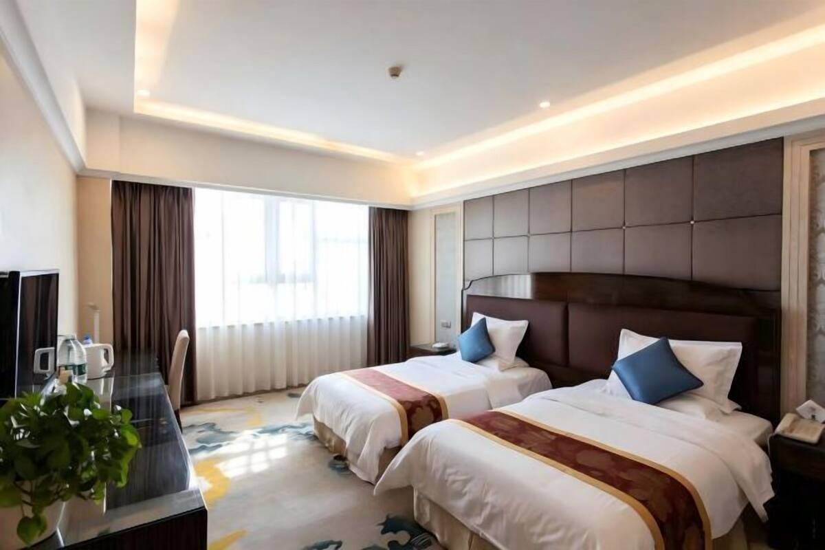A hotel room with two beds, a desk with a chair, a plant, and a large window.