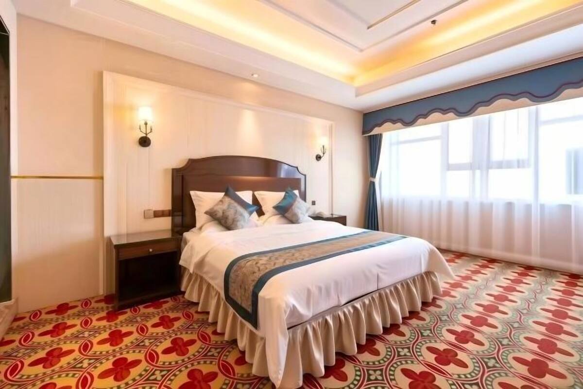 A hotel room with a large bed, a nightstand, a wall-mounted lamp, and a patterned carpet.