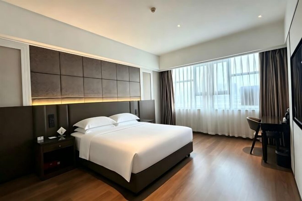 A modern hotel room with a large bed, a bedside table, a chair, and a view of the outdoors through the window.