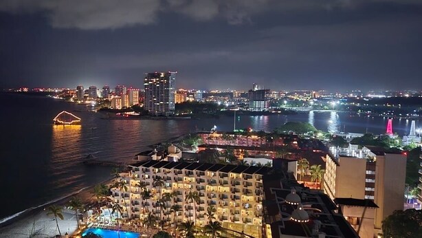 Un paisaje urbano costero nocturno con edificios iluminados, una playa y un hotel con piscina.