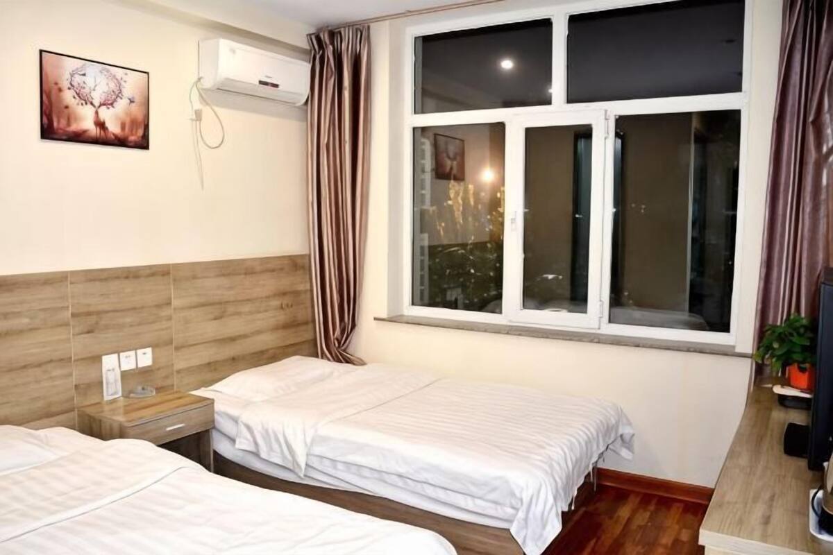 Una habitación de hotel con dos camas, una ventana con cortinas, un aire acondicionado instalado en la pared y una mesita con lámpara.